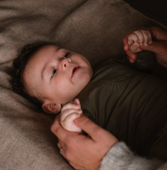Close-up van een baby in een kaki romper met een leeuwenprint, die de hand van een volwassene vasthoudt.