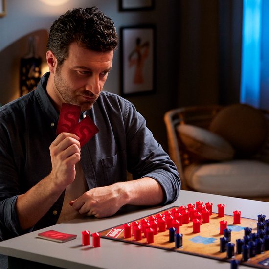 Een man kijkt geconcentreerd naar Stratego-spelkaarten, met het speelbord en rode en blauwe pionnen voor hem.