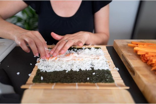 Een persoon maakt sushi met rijst, zeewier en vis op een bamboemat.