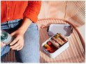 Personne en chemise orange et jean, assise, avec une boîte à lunch grise contenant un sandwich et des légumes.