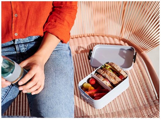 Personne en chemise orange et jean, assise, avec une boîte à lunch grise contenant un sandwich et des légumes.