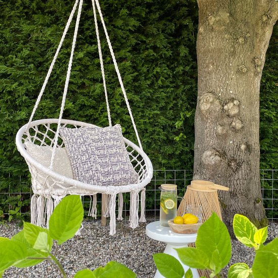 Gebroken wit macramé hangstoel met twee kussens, naast een boom.