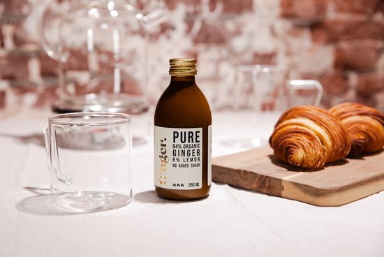 Bruine fles G'nger biologisch gembersap, 94% gember, 6% citroen, zonder toegevoegde suiker, naast een leeg glas en twee croissants op een houten plank.