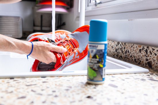 Hand wast oranje en grijze sportschoenen onder de kraan, met een fles Nikwax.