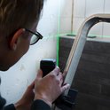 Man met bril stelt een VONROC kruislijnlaser af op een ladder met groene laserlijnen.