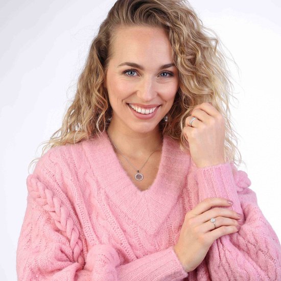 Femme blonde souriante avec des cheveux bouclés, portant un pull rose. Porte une bague et un collier argentés.