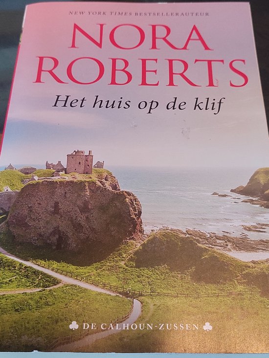 Boekomslag 'Het huis op de klif' van Nora Roberts met een kasteelruïne op een rotsachtige kustlijn.