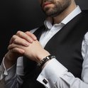 Gevlochten zwarte armband met zilverkleurige details, gedragen door een man in wit overhemd en vest.