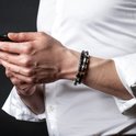 Gevlochten zwarte armband met zilverkleurige details, gedragen door een man in wit overhemd, houdt telefoon vast.