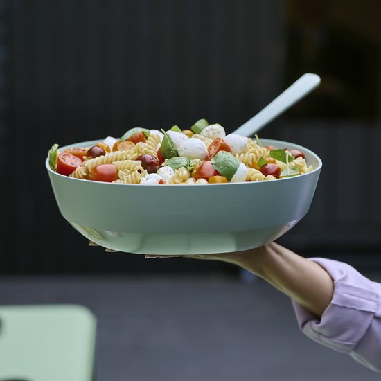 Een hand houdt een grote, lichtgroene kom vol met pastasalade, mozzarella en cherrytomaatjes.