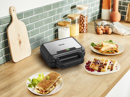 Gesloten Tefal wafelijzer met een tosti en wafels op borden op een houten aanrecht.