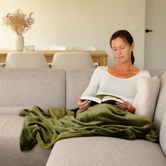 Vrouw in een witte top, onder een olijfgroene plaid, lezend op een beige bank.