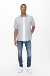Man met korte mouwen in lichtgrijs, open overhemd over een wit T-shirt en distressed jeans.