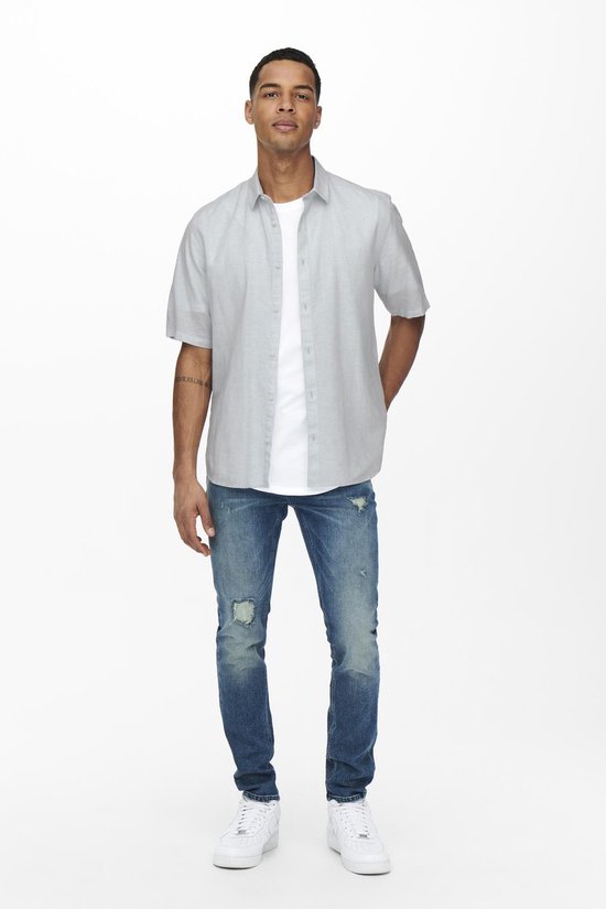 Man met korte mouwen in lichtgrijs, open overhemd over een wit T-shirt en distressed jeans.
