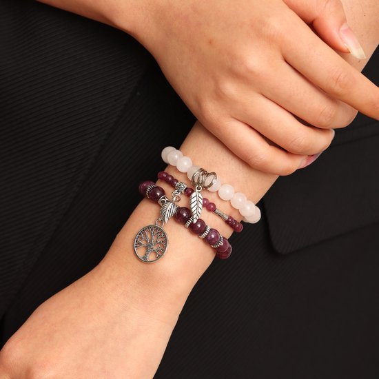 Drie gestapelde armbanden om een arm, met paarse en witte kralen, zilveren bedeltjes van een boom en bladeren.