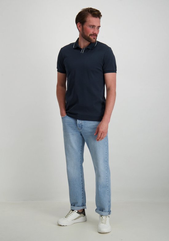 Man met donkerblauw poloshirt en lichtblauwe jeans, met opgerolde pijpen en witte sneakers.