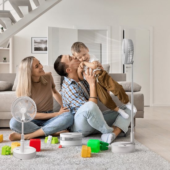 Een gezin geniet van de zomerse koelte van een witte staande ventilator op de vloer, met een tafelventilator en speelgoed op de achtergrond.