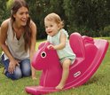 Een baby op een roze Little Tikes hobbelpaard in het gras, met een lachende vrouw op de achtergrond.
