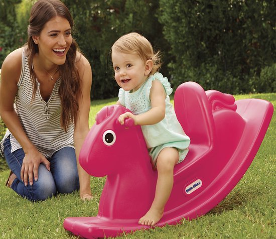 Een baby op een roze Little Tikes hobbelpaard in het gras, met een lachende vrouw op de achtergrond.