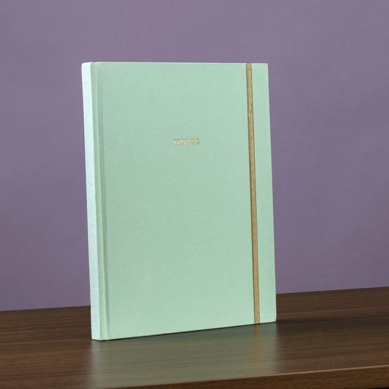 Lichtgroen notitieboek met de tekst 'NOTES' in goudfolie en een gouden elastische band, staand op een houten tafel.