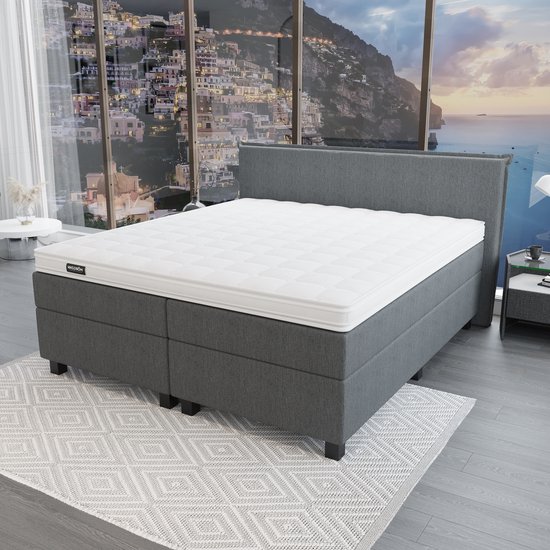 Grijze boxspring met wit gestreept matras en een "Mörgenn" logo, voor een raam met uitzicht op een kuststad.