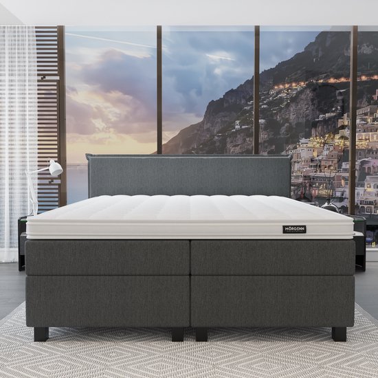 Grijze boxspring met wit matras, oranje plaid, gele kussens en uitzicht op een kuststad.