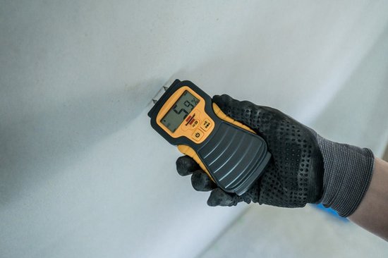 Hand met zwarte handschoen die een gele en grijze vochtmeter vasthoudt tegen een witte muur. Het display toont 5,9%.