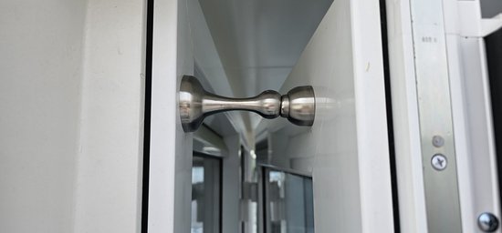 Een zilverkleurige, gebogen deurstopper met een ronde magneet aan het uiteinde, gemonteerd op een witte deurpost.