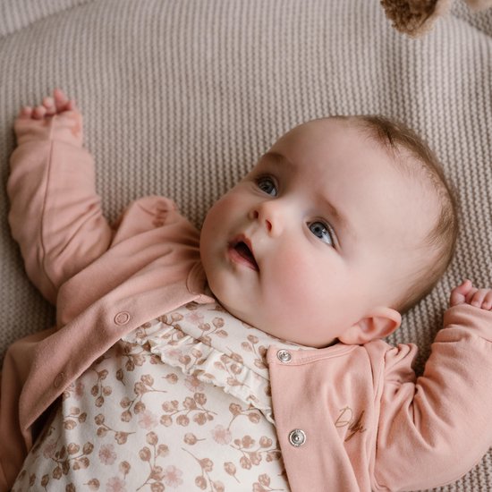 Baby met grote blauwe ogen ligt op een beige plaid en draagt een roze vestje over een wit jurkje met bloemenprint.