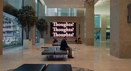 Man zit in een moderne lobby, kijkt naar een scherm met 'Thoughts? Thoughts? Thoughts?'.