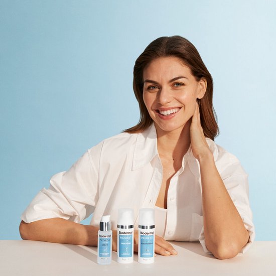 Een lachende vrouw met bruin haar draagt een wit shirt. Drie Biodermal producten staan voor haar op tafel: een serum en twee crèmes.
