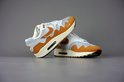 Twee witte met oranje Nike Air Max 1 Patta sneakers met beige en bruine zolen, op een grijze achtergrond.