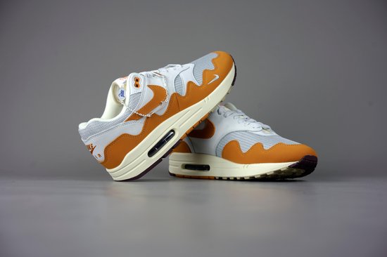 Twee witte met oranje Nike Air Max 1 Patta sneakers met beige en bruine zolen, op een grijze achtergrond.