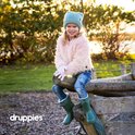 Lachend meisje met blonde haren, zittend op een boomstam in een park. Draagt een roze jas, spijkerbroek en groene laarzen.