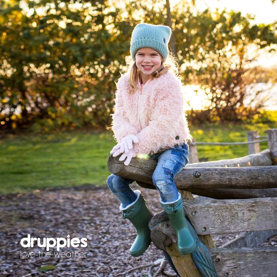 Lachend meisje met blonde haren, zittend op een boomstam in een park. Draagt een roze jas, spijkerbroek en groene laarzen.