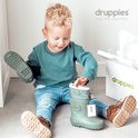 Jongetje zit op de grond, speelt met groene laarzen en bruine laarsjes. Een doos van Druppies staat ernaast.