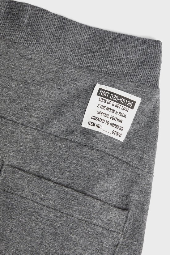 Close-up van het label op een donkergrijze joggingbroek met tekst: 'NMT 028-65196 LOOK UP & GET LOST 2 THE MOON & BACK SPECIAL EDITION CREATED TO IMPRESS ITEM NO.: 028/6'.