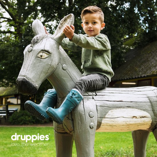 Jongen met groene trui zit op een houten ezel, draagt petrolblauwe regenlaarzen.
