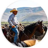 PVC Schuimplaat Muurcirkel - Cowboy op zijn Paard tussen Kudde bij Bergen - 90x90 cm Foto op Muurcirkel (met ophangsysteem)