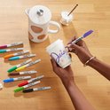 Een hand met paarse Sharpie stift kleurt de tekst 'Daisy' op een witte mok. Diverse kleuren stiften liggen ernaast.