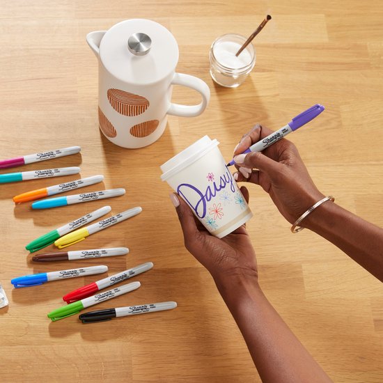 Een hand met paarse Sharpie stift kleurt de tekst 'Daisy' op een witte mok. Diverse kleuren stiften liggen ernaast.