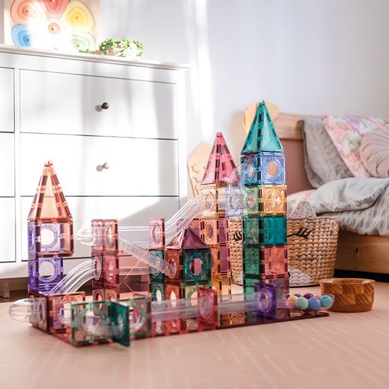 Een torenhoge baan van gekleurde Connetix-tegels en transparante buizen, gebouwd in een kinderkamer met een bed en mand.
