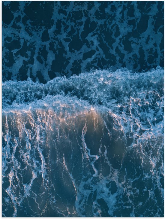 Bovenaanzicht van oceaangolven in blauwe tinten met wit schuim en een zachte roze gloed.