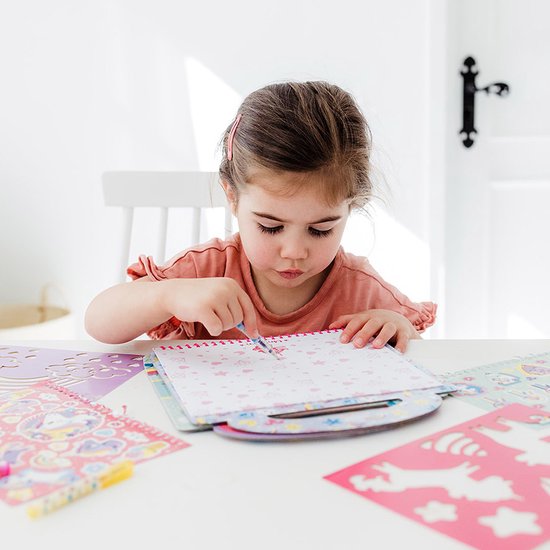 Une jeune fille en chemise rose dessine dans un livre d'activités Mini MyMy avec des animaux mignons et des étoiles.