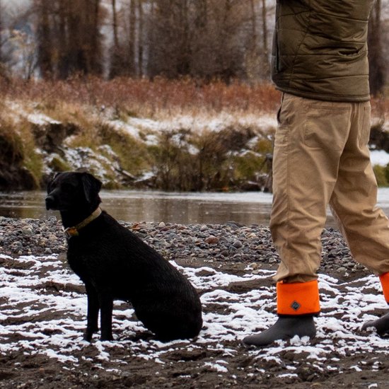 Persoon draagt Woody Max laarzen met omgeslagen oranje rand naast een hond op een besneeuwde ondergrond.