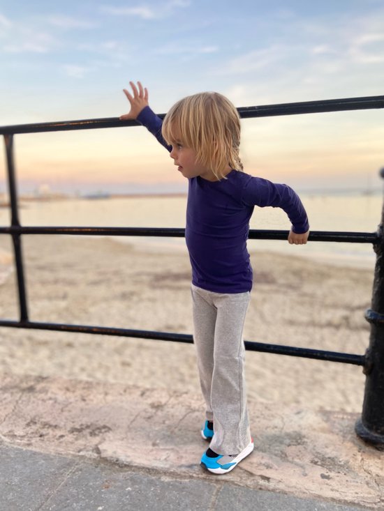 Een jongen met blond haar, gekleed in een donkerblauwe top en een grijze wijde broek, staat bij een hek aan zee.