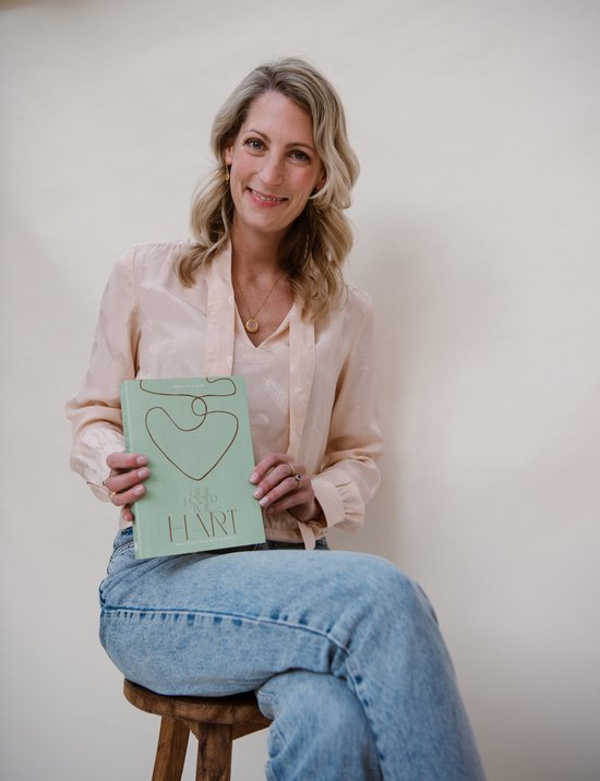 Vrouw met blonde haren in een lichtroze blouse en spijkerbroek, zittend op een krukje en een groen boek vasthoudend met de tekst 'UIT JE HOOFD IN JE HART'.