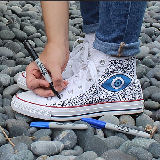 Een persoon tekent met een zwarte Sharpie marker op een witte hoge sneaker, die versierd is met een blauw oog.
