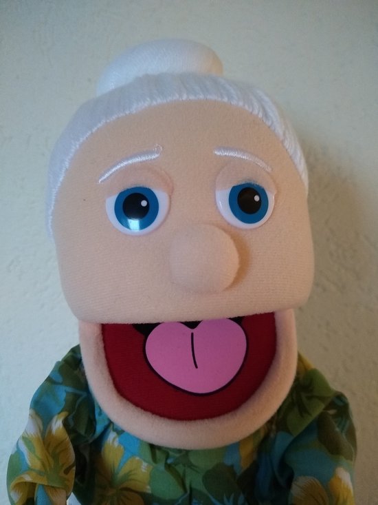 Gezicht van een handpop van een oma met blauwe ogen en open mond met roze tong. Witte haren in een knot, draagt groen met bloemenjurk en schort.