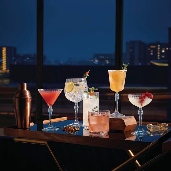 Een barwagen met diverse cocktails, een koperen shaker en een gebogen schaar, met uitzicht op een nachtelijke stad.
